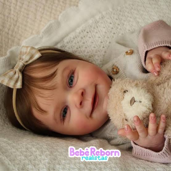 Bebê Reborn sorridente com laço no cabelo e ursinho de pelúcia